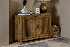 The Yasha Reeded Mango Wood Sideboard by nkuku, with vertical grooves, stands against a beige wall. On top are a vase with dried flowers, a book, candle, framed photo, and brass lamp. A woven basket sits beside a blue and beige rug.