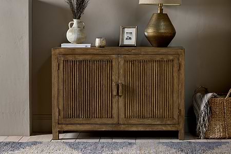 The nkuku Yasha Reeded Mango Wood Sideboard stands against a beige wall, topped with a ceramic vase of dried flowers, candle, framed photo, and gold lamp. A woven basket sits on the floor nearby, with part of a rug visible.