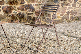 A brown nkuku Yealme Iron Bistro Chair with a slatted seat and backrest stands on gravel before a stone wall of irregular brown and tan rocks. Another piece of industrial outdoor furniture is partially visible at the left edge.