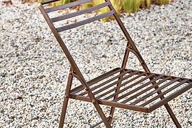 A close-up of the nkuku Yealme Iron Bistro Chair, an industrial outdoor piece featuring brown metal slats on the seat and backrest, sits on gravel with blurred green plants in the background.