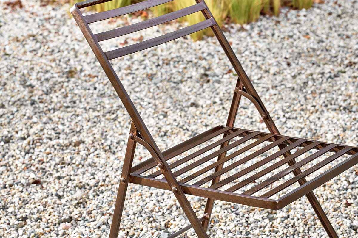 A close-up of the nkuku Yealme Iron Bistro Chair, an industrial outdoor piece featuring brown metal slats on the seat and backrest, sits on gravel with blurred green plants in the background.