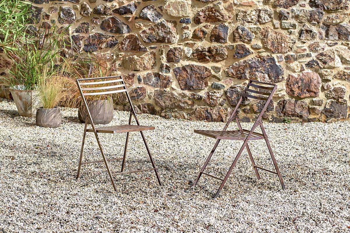 Two brown nkuku Yealme Iron Bistro Chairs face each other on a gravel patio, set against a rustic stone wall and surrounded by potted plants, creating a relaxed spot to enjoy industrial outdoor furniture in the fresh air.