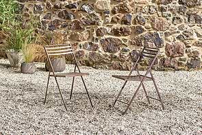 Two brown nkuku Yealme Iron Bistro Chairs face each other on a gravel patio, set against a rustic stone wall and surrounded by potted plants, creating a relaxed spot to enjoy industrial outdoor furniture in the fresh air.