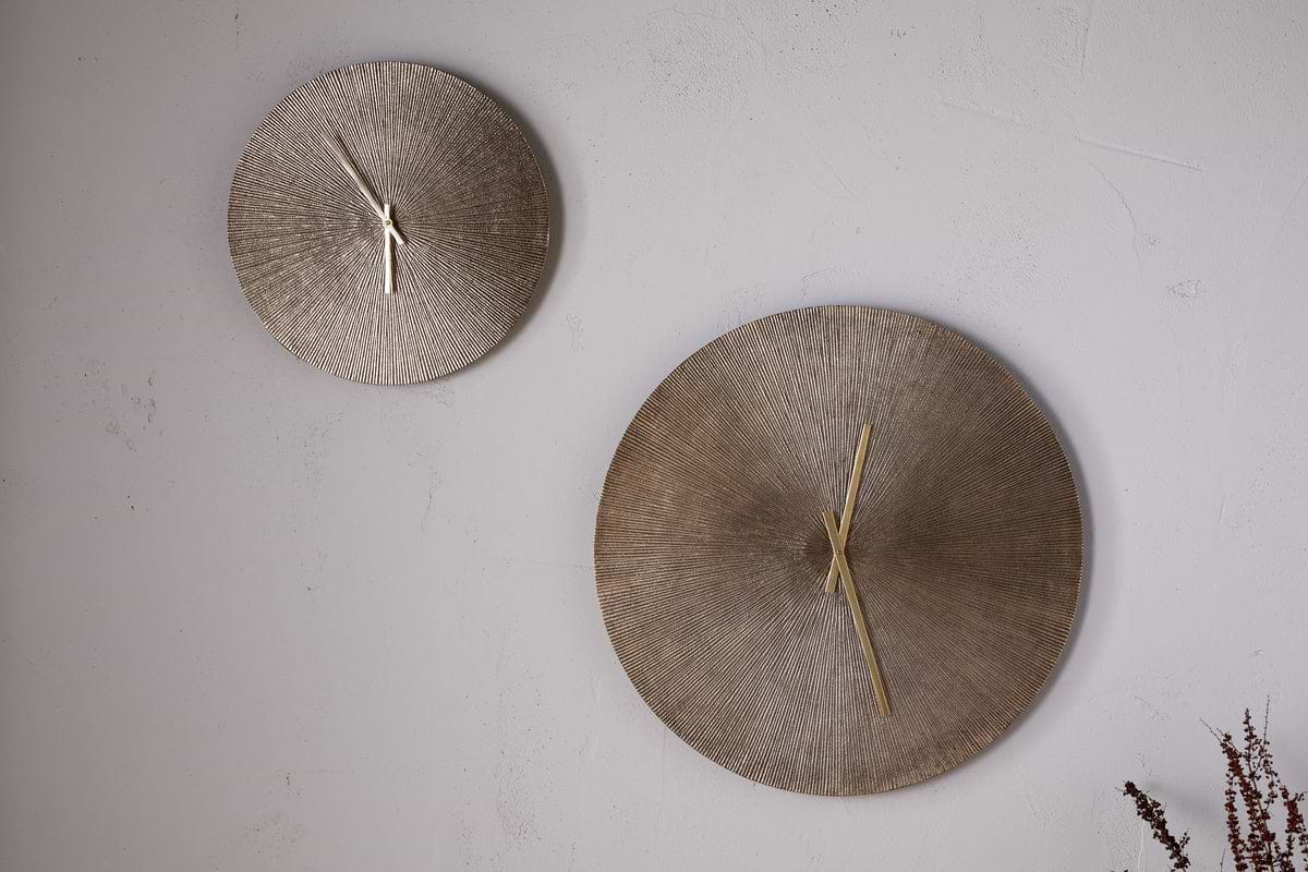 Two Zuwena Etched Wall Clocks by nkuku in antique brass are mounted on a light textured wall. The minimalist display features different sizes, with dried plant stems in the lower right corner adding subtle decor.