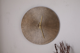 The nkuku Zuwena Etched Wall Clock in Antique Brass, featuring a textured radial gold face and minimal gold hands, hangs on a plain light wall with dried brown plant stems in the corner for a minimalist accent.