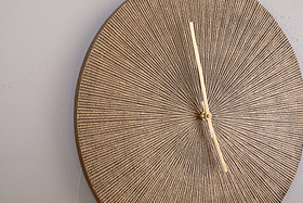 The nkuku Zuwena Etched Wall Clock in Antique Brass features a textured, round face with a radial beige pattern and slim pale gold hands. This minimalist, unmarked clock adds sophistication when mounted on a light gray wall, displaying 12:25.