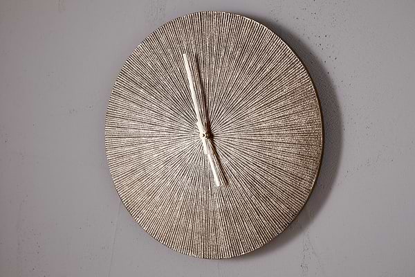 The Zuwena Etched Wall Clock by nkuku features a radial, antique brass finish and slim silver hands set to 11:00. This minimalist piece has no numbers and is mounted on a plain light gray wall.
