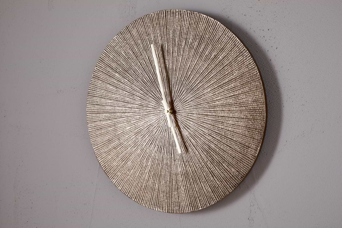 The Zuwena Etched Wall Clock by nkuku features a radial, antique brass finish and slim silver hands set to 11:00. This minimalist piece has no numbers and is mounted on a plain light gray wall.