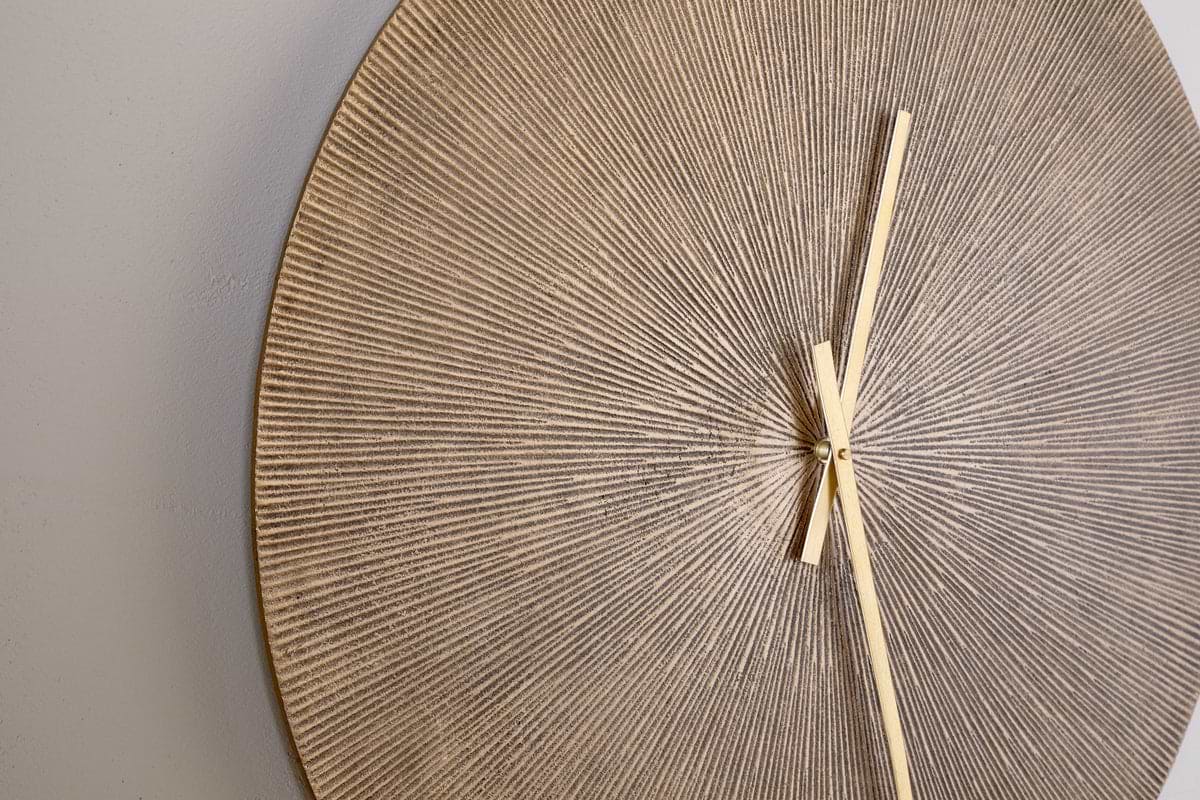 A close-up of the nkuku Zuwena Etched Wall Clock - Antique Brass shows its round, textured face with a beige and gold radial pattern. Minimalist gold hands point to 1:25 against a light gray background, with part of the clock face visible.