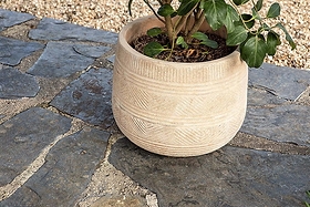 A leafy green plant sits in nkuku’s Zadie Etched Ceramic Planter (Neutral, Set of 2), featuring subtle geometric patterns. The handmade ceramic pot rests on blue-gray flagstones with tan pebbles in the background.