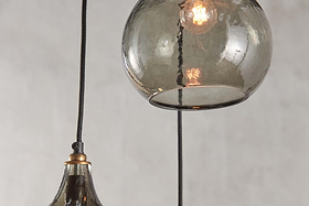 Close-up of the nkuku Ziva Recycled Glass Cluster Pendant in Green Smoke, featuring two hanging fixtures—one globe and one bell-shaped. Both are suspended by black cords with warm bulbs glowing against a neutral gray background.