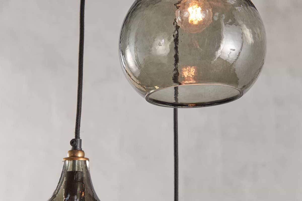Close-up of the nkuku Ziva Recycled Glass Cluster Pendant in Green Smoke, featuring two hanging fixtures—one globe and one bell-shaped. Both are suspended by black cords with warm bulbs glowing against a neutral gray background.