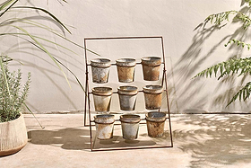 The nkuku Abari Planter Stand displays nine aged zinc pots in a 3x3 grid. Set on concrete by a beige wall, it's framed by leafy plants and gentle shadows, bringing rustic garden style and sunlit charm to any space.