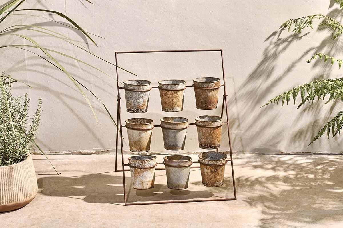 The nkuku Abari Planter Stand displays nine aged zinc pots in a 3x3 grid. Set on concrete by a beige wall, it's framed by leafy plants and gentle shadows, bringing rustic garden style and sunlit charm to any space.