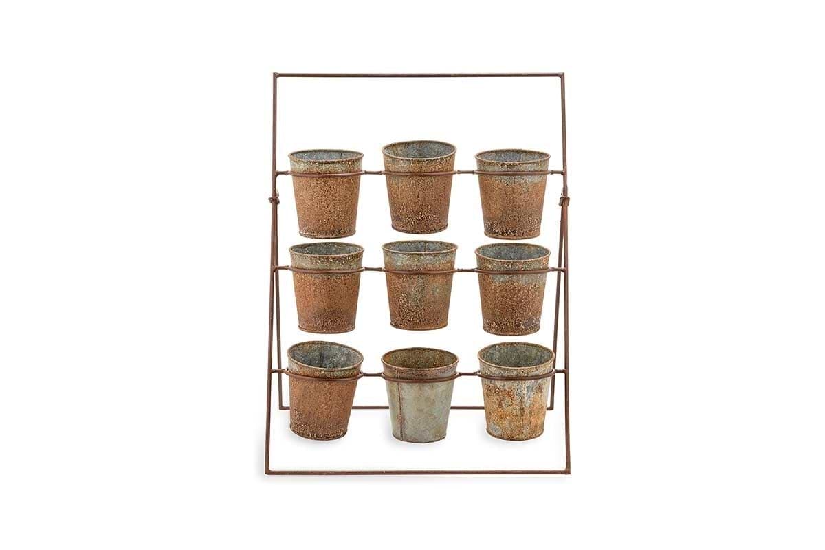 The Abari Planter Stand by nkuku holds nine rustic aged zinc pots in three rows. Angled backward for display, this minimalist metal rack showcases weathered pots—ideal as unique garden decor—against a plain white background.