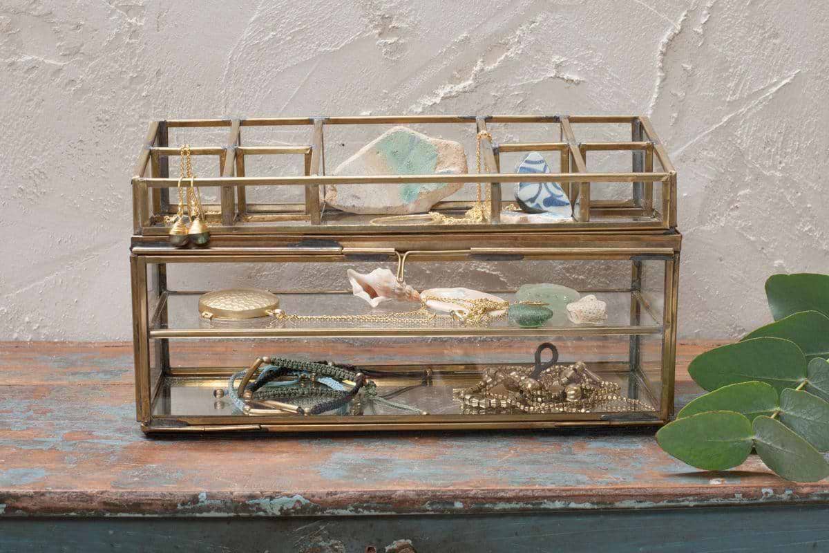 The Bequai Open Jewellery Box by nkuku features two glass and brass levels for storing necklaces, bracelets, stones, or seashells. It rests on a blue wooden surface with eucalyptus leaves nearby and a textured beige wall in the background.