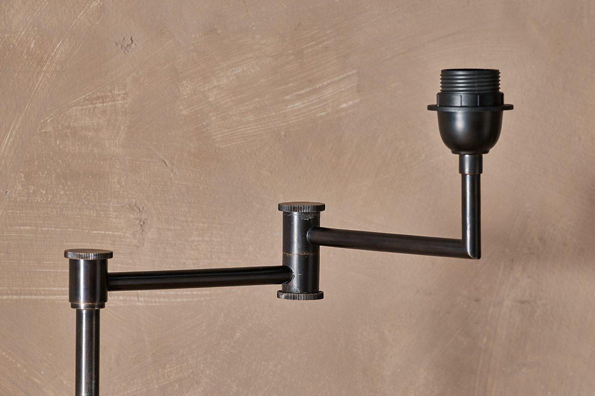 Close-up of the Kara Iron Floor Lamp - Bronze by nkuku, showing its aged bronze adjustable arm with bulb socket against a textured beige wall. Minimalist and industrial in design; bulb and jute lampshade not included.