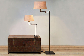 A Kara Iron Floor Lamp - Bronze by nkuku stands on the wooden floor near a pale wall, beside a matching table lamp with beige or jute shade and black or aged bronze base; the smaller rests atop an old wooden trunk.