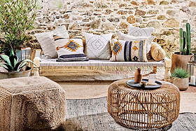 A cozy, rustic seating area showcases the nkuku Kutumba Mango Day Bed with patterned cushions, a handwoven jute pouf, a round wicker coffee table topped with pottery, and potted plants against a stone wall for a boho, natural vibe.