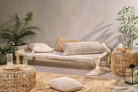 A minimalist room features the nkuku Kutumba Mango Day Bed with neutral pillows and throw, woven rattan side tables, a handwoven jute seat, potted plants, candles, and a lantern for a cozy, natural feel.