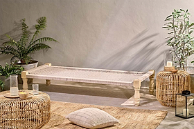 The nkuku Kutumba Mango Day Bed with a handwoven jute seat and light wood legs sits against a beige wall, surrounded by potted plants, a rattan table, ottoman, candle, lantern, and beige cushion on a patterned rug.