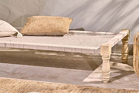 The Kutumba Mango Day Bed by nkuku, with light wooden legs, sits on a concrete floor next to a textured beige rug. Two woven cushions—one cream, one brown—rest on the Indian day bed as sunlight softens the pale wall behind.