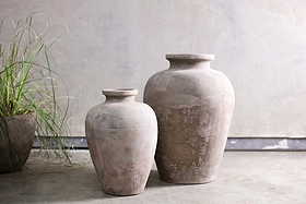 Two nkuku Affiti Clay Tapered Pots - Large, with a rustic handmade finish, stand side by side on a concrete floor against a muted wall. To the left, long thin leaves from a potted plant partially enter the frame.