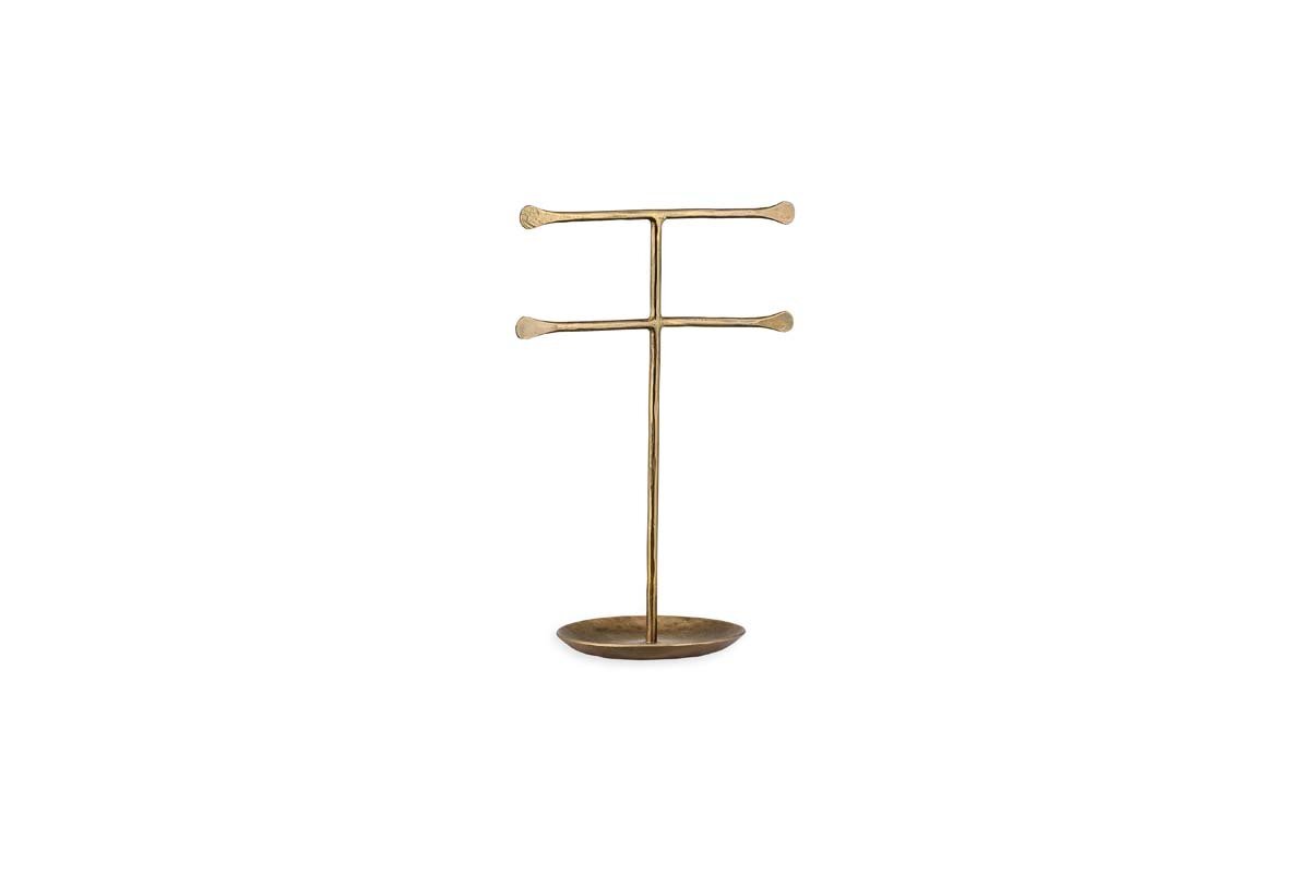 The Liman Jewellery Stand by nkuku is a minimalist brass stand with a matte finish, featuring a vertical rod and two horizontal bars for jewelry display. It has an oval base that serves as a ring tray, all set against a plain white background.