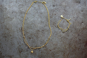 The nkuku Mai Necklace, featuring two gold chains with small beads, pyrite stones, and a round pendant, is shown on a textured gray surface beside a matching bracelet with pyrite beads and a charm-accented clasp.
