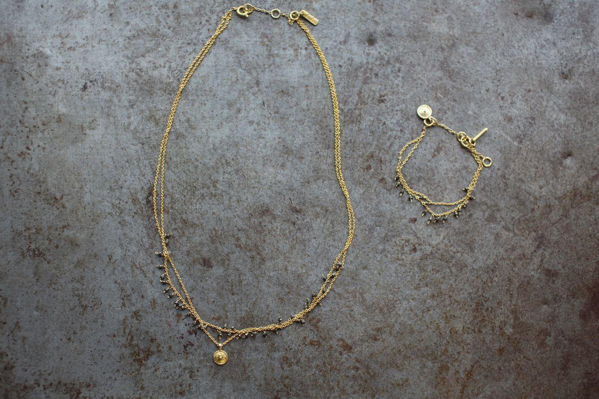 The nkuku Mai Necklace, featuring two gold chains with small beads, pyrite stones, and a round pendant, is shown on a textured gray surface beside a matching bracelet with pyrite beads and a charm-accented clasp.