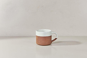 The Mali Ribbed Coffee Mug - White (Set of 2) by nkuku features a handmade design with a white upper half, ribbed terracotta lower half, and a white handle, displayed on a light beige surface against a pale background.