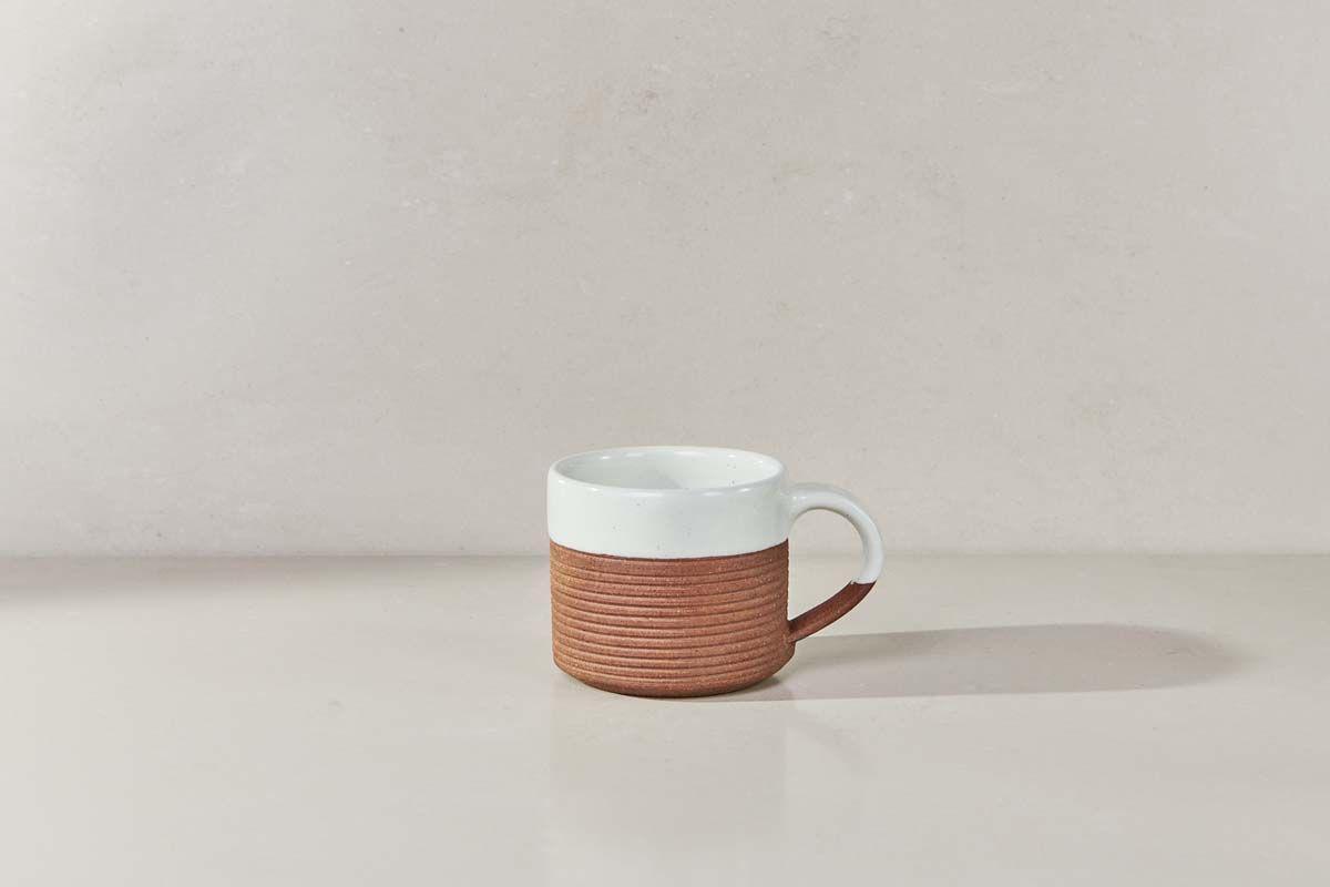 The Mali Ribbed Coffee Mug - White (Set of 2) by nkuku features a handmade design with a white upper half, ribbed terracotta lower half, and a white handle, displayed on a light beige surface against a pale background.
