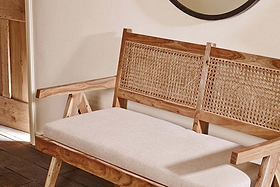 The Nalrina acacia and cane sofa by nkuku, with a cream linen seat pad, sits against a white wall near a wooden door frame. A round mirror is partially visible in the upper right corner.