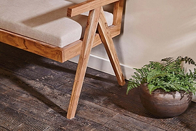 The Nalrina acacia and cane sofa by nkuku rests on a dark wood floor beside a potted fern, featuring a beige linen seat pad.