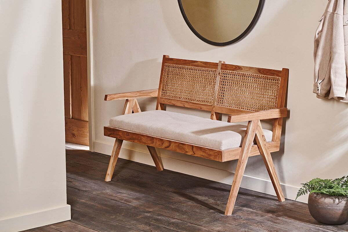 The nkuku Nalrina acacia and cane sofa with a beige cushion sits against a light wall, complemented by a round mirror above, a potted plant nearby, and a coat to the side. The dark wood floor adds warmth to the space.