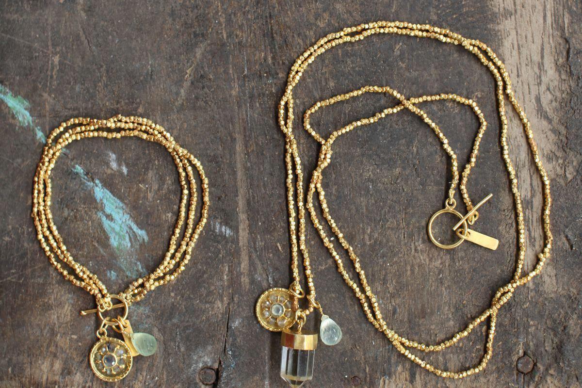 Two sets of gold-plated, handmade jewelry from nkuku rest on a rustic wooden surface. On the left is the Pandita Charm Bracelet with a gear, crystal bead, and round pendant; on the right is a necklace with similar charms and a vial-shaped pendant.
