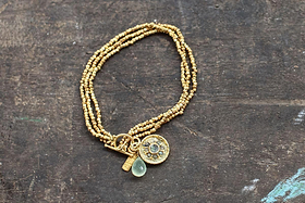 The Pandita Charm Bracelet by nkuku is a handmade piece with three strands of small gold-plated beads. It features an ornate medallion, a pale green teardrop stone, and a gold bar charm, set on a dark wooden surface.