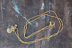 The Pandita Charm Necklace by nkuku is a handmade gold beaded necklace with a toggle clasp, crystal quartz, an intricately designed round pendant, and a small bead, displayed on weathered dark wood.