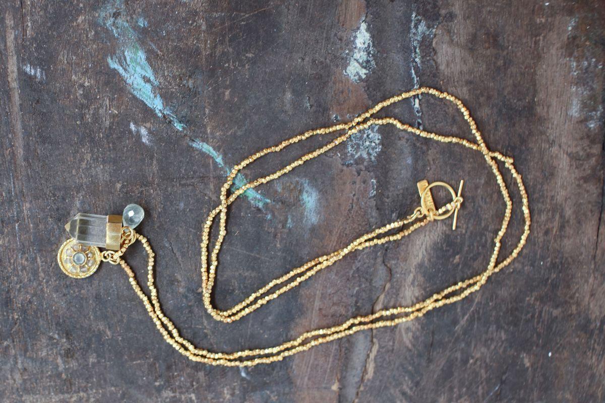 The Pandita Charm Necklace by nkuku is a handmade gold beaded necklace with a toggle clasp, crystal quartz, an intricately designed round pendant, and a small bead, displayed on weathered dark wood.