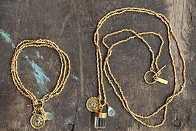 The Pandita Charm Necklace by nkuku sits on a rustic wooden surface, paired with a gold beaded bracelet. Each handmade piece features round medallions and crystal quartz beads, highlighting the collection’s simple yet elegant design.