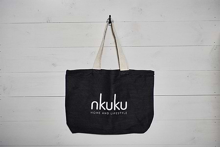 The nkuku Jute & Cotton Tote Bag, a large black handmade-style tote with sturdy beige handles and "nkuku HOME AND LIFESTYLE" in white on the front, hangs on a hook against a white wooden wall.