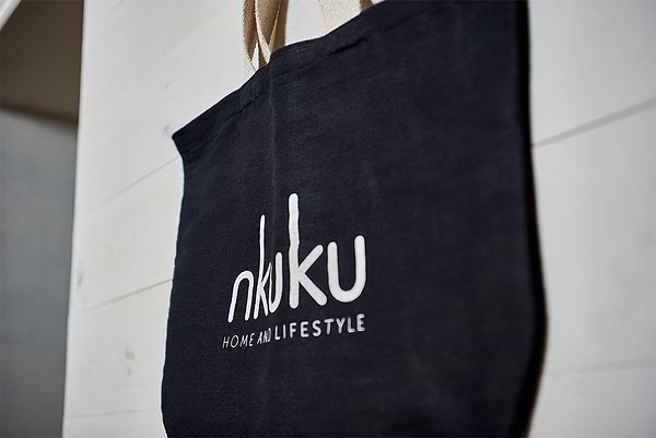 The Jute & Cotton Tote Bag by nkuku, in black with beige handles, hangs on a white wooden wall. The bag displays the white nkuku logo in lowercase, with "HOME AND LIFESTYLE" below it in small uppercase letters.