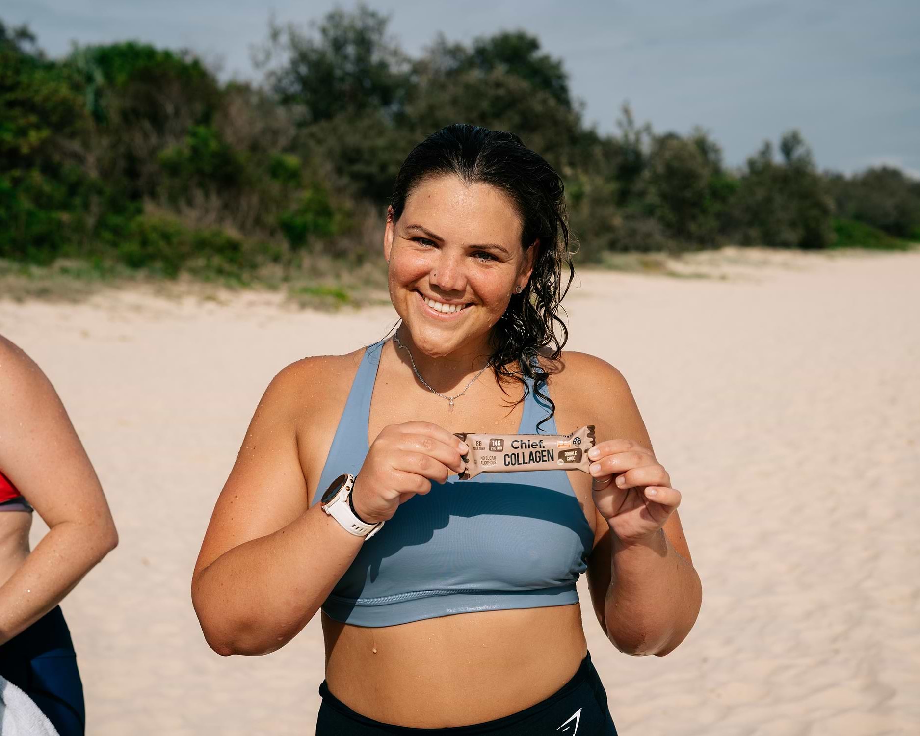A woman on the beach in activewear smiling at the camera and holding up a Chief Collagen Bar