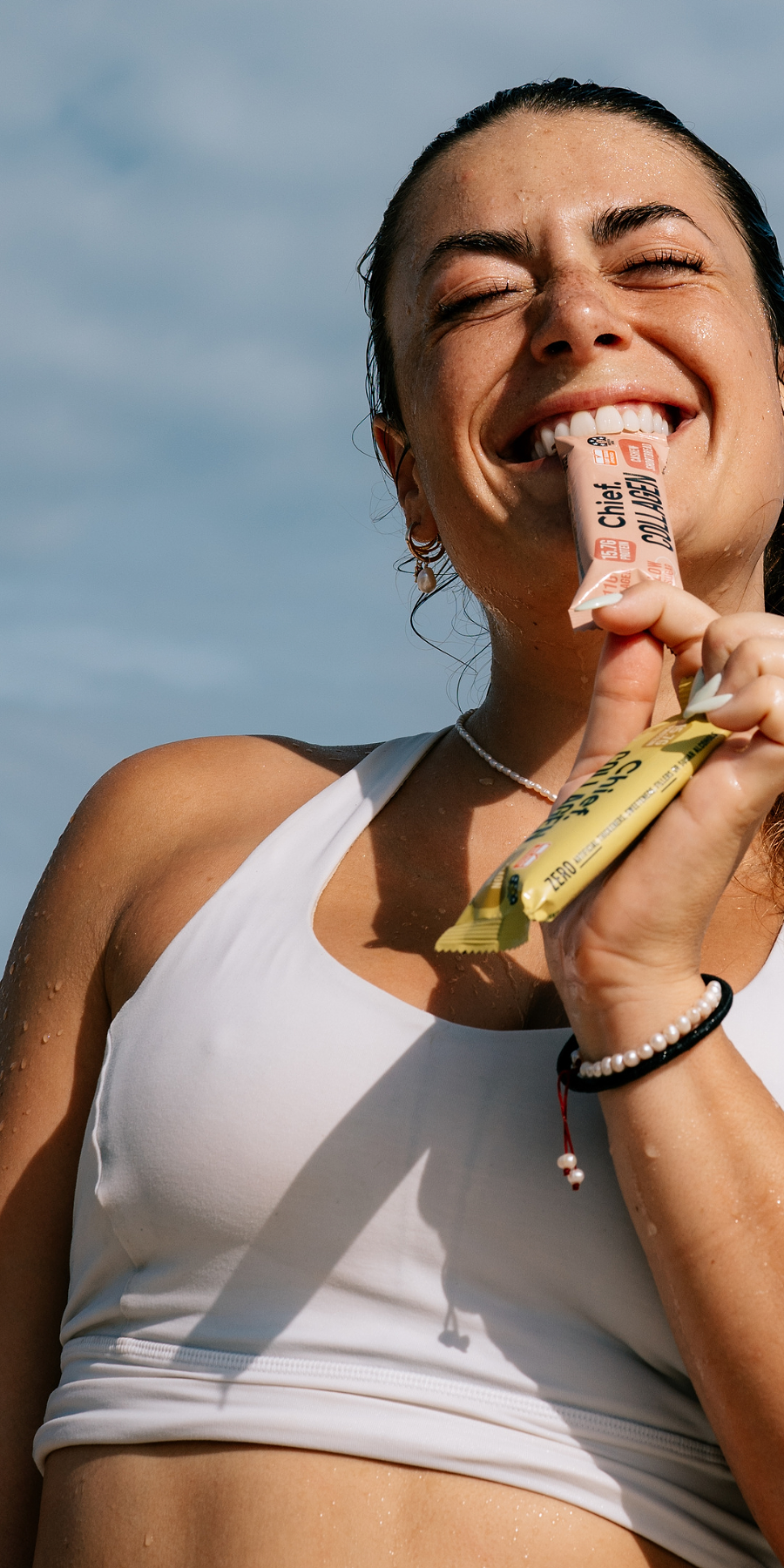 A woman outdoors wearing a white sports bra, smiling as she holds a pink protein powder tube in her teeth and a yellow snack bar in her hand against a blue sky background.