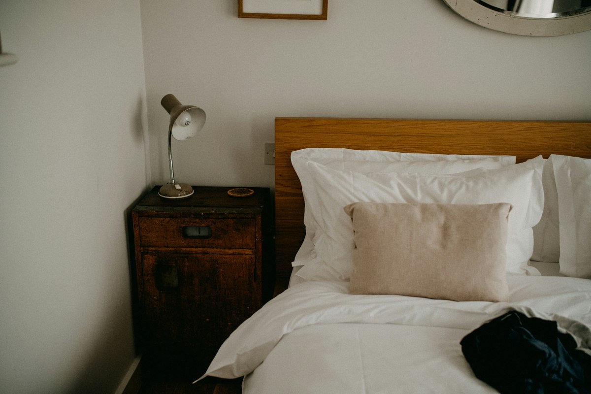 A bed with white bedding, a beige cushion, and a wooden bedside table.