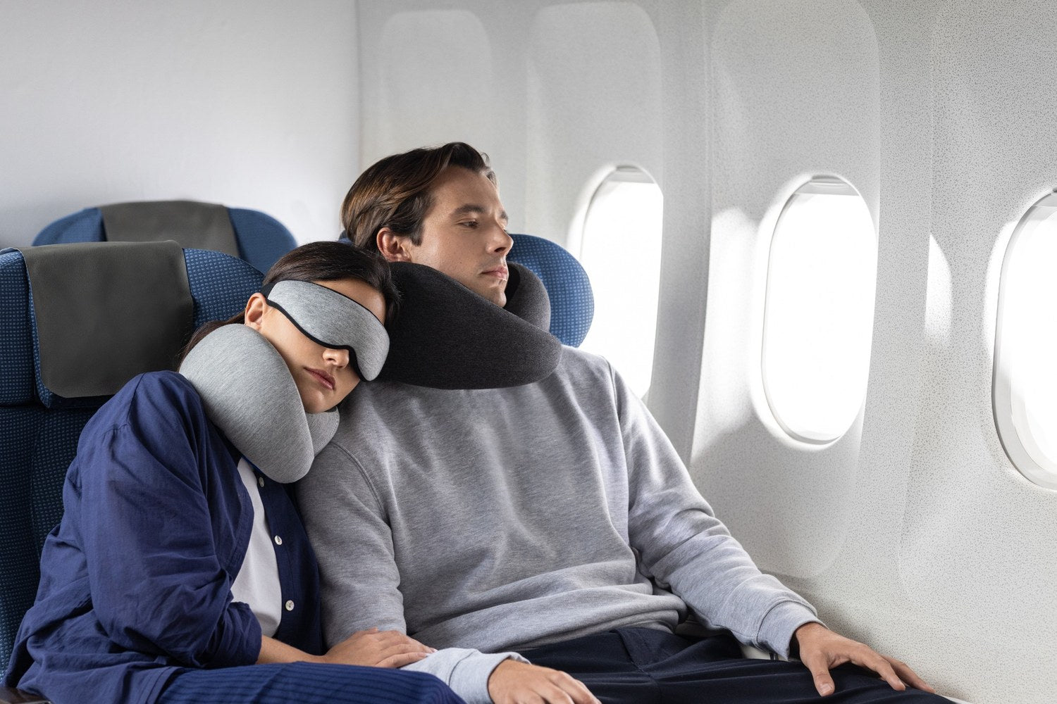 Two travelers seated on a plane wearing Go neck pillows, with one resting in an eye mask.