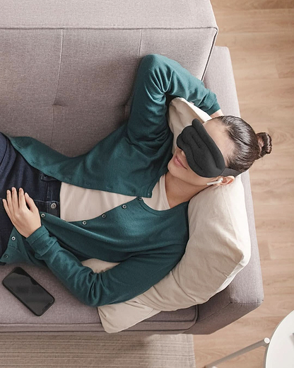 High-angle view of a woman lying comfortably on a grey fabric couch wearing a green cardigan and the cushioned Hot and Cold Eye Mask in Midnight Grey.