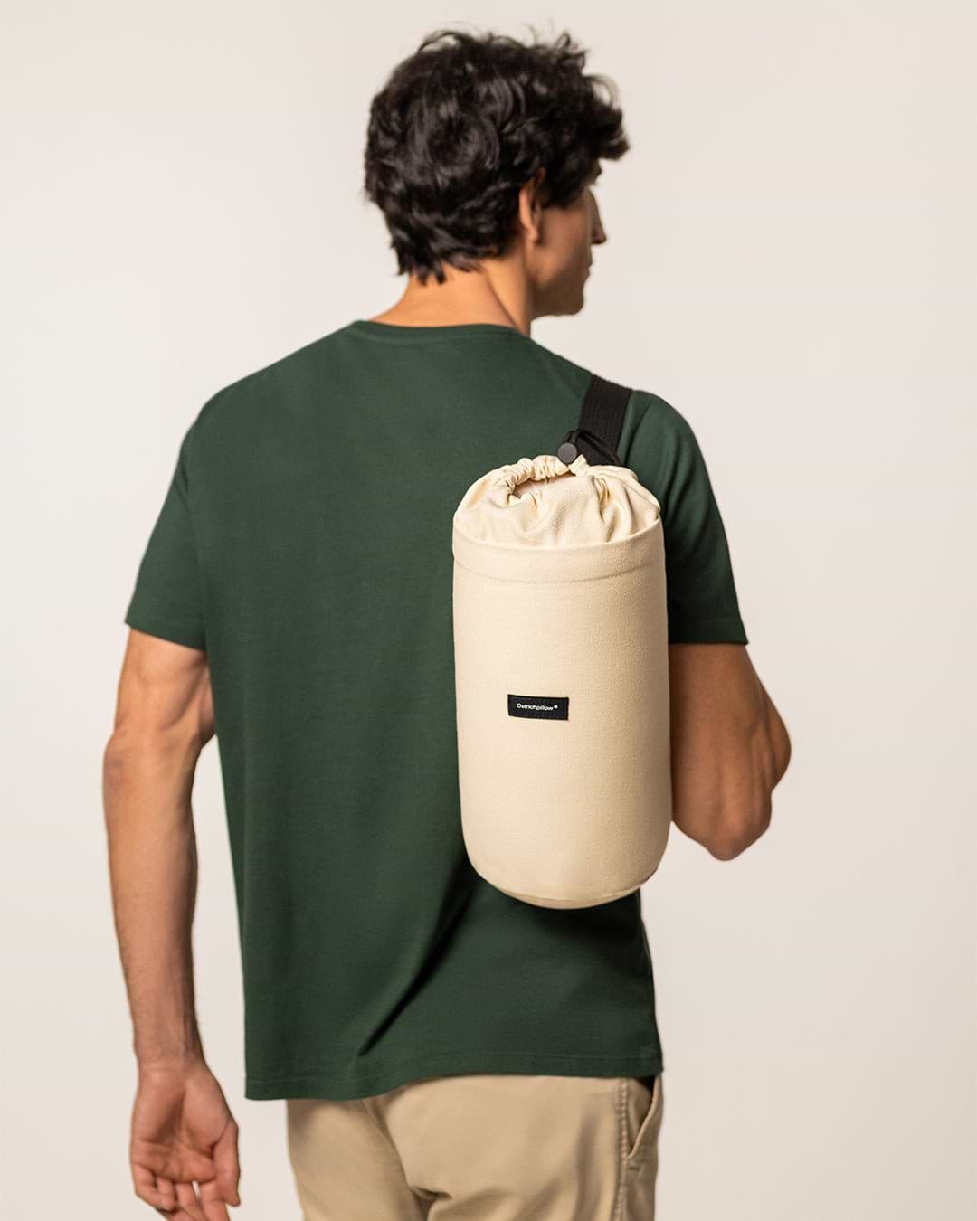 Back view of a man carrying a rolled-up Ostrichpillow Bed Pillow inside its cream-colored drawstring travel bag with a black shoulder strap.