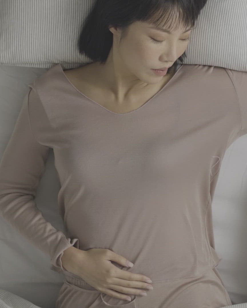 A woman lying in bed places Heatbag onto her lower abdomen, gently resting her hands over it for soothing warmth and comfort.