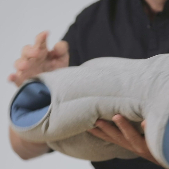 A man demonstrates how to wear Original Napping Pillow. He pulls it over his head and ensures his nose and mouth align with the breathing opening.